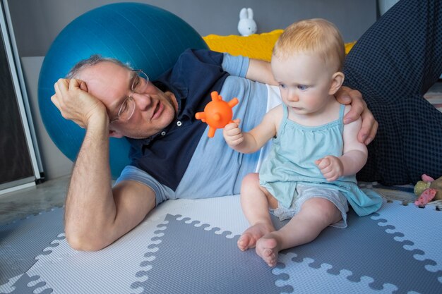 Adorable red-haired newborn sitting on floor and playing toy. Happy grandfather in eyeglasses and blue shirt lying near grandchild and telling story. Family, infancy and childhood concept