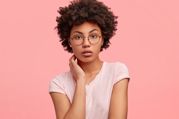 Adorable serious African American female with self assured expression, keeps hands on neck, dressed in casual shirt in one tone with wall, has healthy skin, models indoor. People and ethnicity