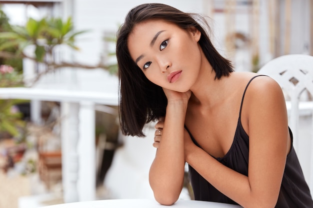 Adorable serious Asian female has serious expression, poses at cafe alone, feels bored in unknown country, dressed casually, has good recreation.