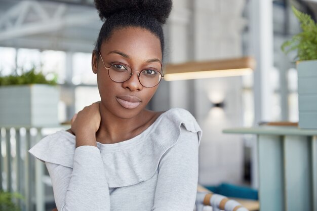 Adorable serious dark skinned female with confident expression, wears glasses, works on scientific report, poses indoor.