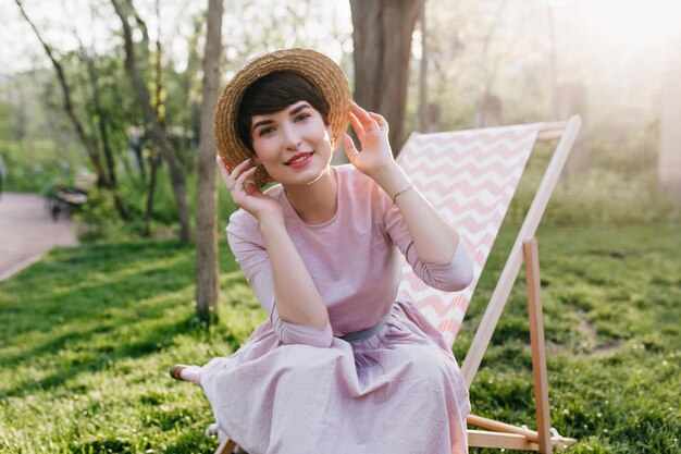 Adorable short-haired lady playfully posing in forest, sitting in comfortable recliner on grass background. Outdoor portrait of cute girl in hat with pale skin enjoying sunshine in weekend.
