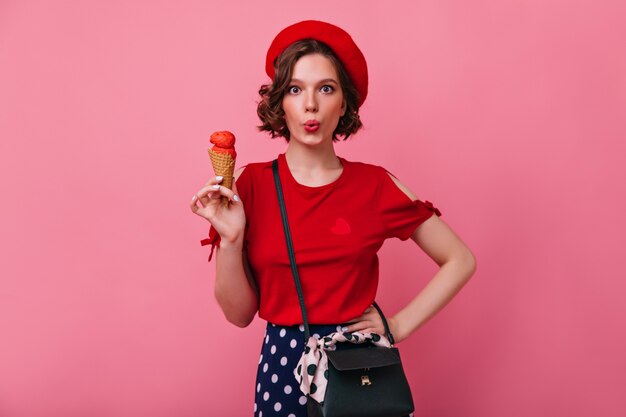 Adorable slim girl in red blouse eating ice cream. Indoor shot of gorgeous caucasian woman with short haircut posing.