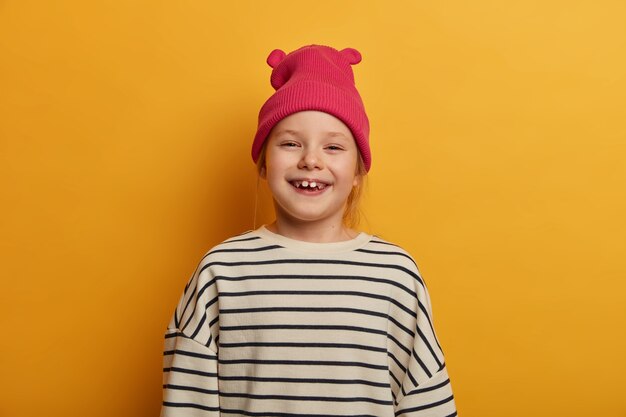 사랑스러운 작은 아이가 긍정적으로 치아를 잃고, 기분이 좋고, 즐거운 하루를 기뻐하고, 분홍색 유행 모자와 줄무늬 느슨한 스웨터를 입고, 노란색 벽에 포즈를 취하고, 기억에 남는 사진을 만듭니다.