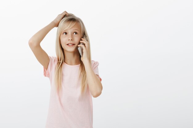 Adorable smart female kid with blond hair, talking on smartphone, scratching head and looking aside