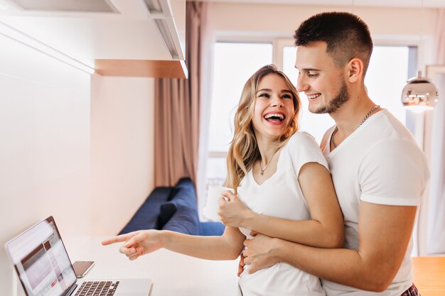 Adorable smiling girl pointing finger to laptop screen. Indoor shot of laughing couple watching funny video.