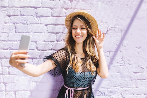 Adorabile ragazza sorridente in cappello di paglia alla moda che fa selfie mentre aspetta un amico fuori