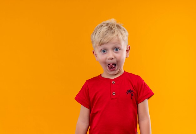 an adorable smiling little boy with blonde hair and blue eyes wearing red t-shirt showing his tongue