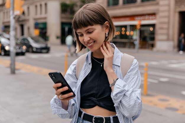 青いシャツと黒いトップを身に着けている街でスマートフォンを保持しながら笑顔の愛らしい笑顔ストレートショートヘアバックパックで暖かい日に街で壁を張っている白人女性の外の写真