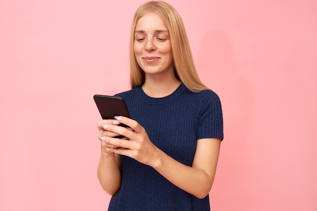 Adorable stylish teenage girl scrolling her newsfeed via social network using high speed internet connection on smart phone