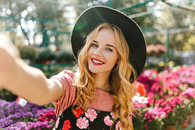 Adorable white woman taking picture of herself in glasshouse with flowers. Laughing pleasant woman making selfie on orangery.