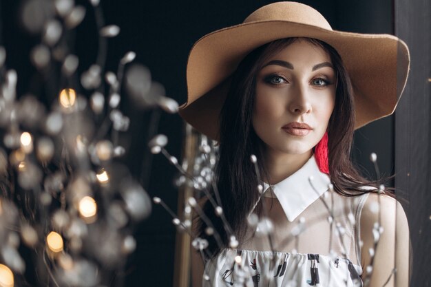 Adorable woman in beige hat and with fashion look poses in the dark studio