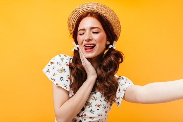 Adorable woman in boater and with bows on her hair shows her tongue and takes selfie on orange background.