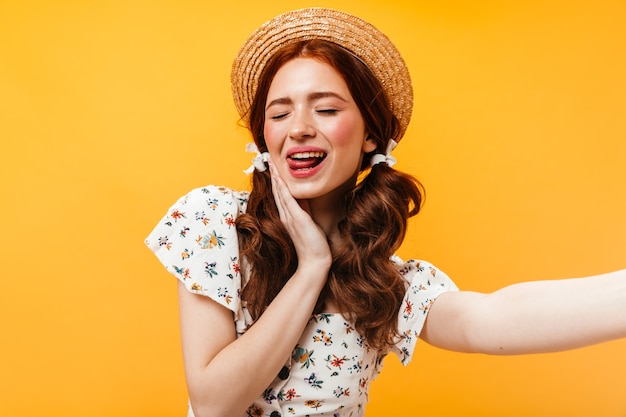 Adorable woman in boater and with bows on her hair shows her tongue and takes selfie on orange background.