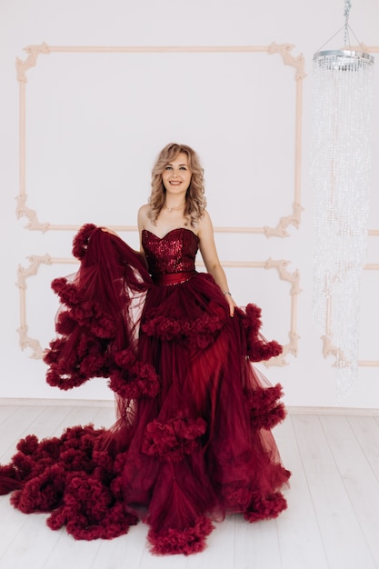 Adorable woman in red burgundi dress poses in a bright luxury room with large chandelier