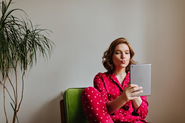 Adorable woman taking selfie with kissing face expression. Indoor shot of curly woman in pajama using digital tablet.