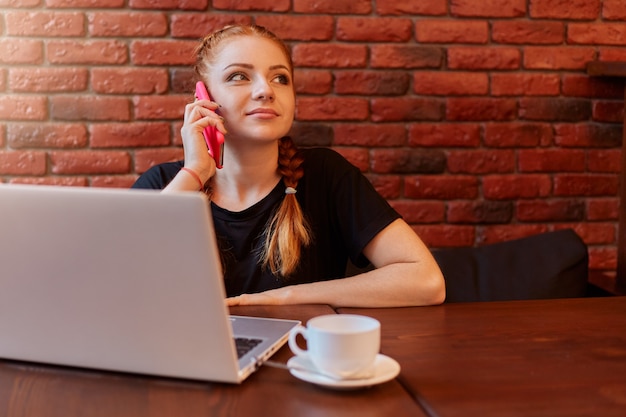 Adorable woman talking on smartphone in cafe while working online via laptop