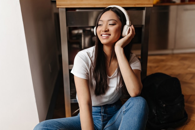 Adorable woman in white headphones posing with smile on floor in kitchen