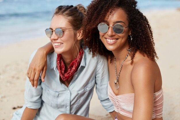 Adorable woman with Afro hairstyle embraces her female partner, begin lesbians, enjoy togetherness and calm atmosphere at seaside, admire views, have happy looks. People, lifestyle, recreation