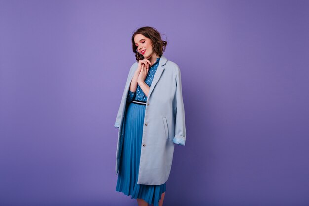 Adorable woman with trendy makeup looking down during photoshoot.  cheerful curly girl in blue coat.