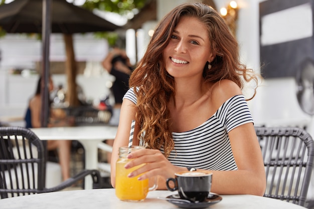 Adorable young female with dark long hair, dressed in striped t- shirt in coffee shop, drinks fresh juice and espresso.