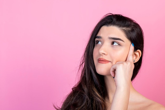 Adorable young girl holding her finger to her head and thinking on pink background High quality photo