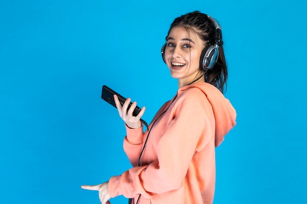 전화를 들고 파란색 배경에 헤드폰을 끼고 있는 사랑스러운 어린 소녀 고품질 사진