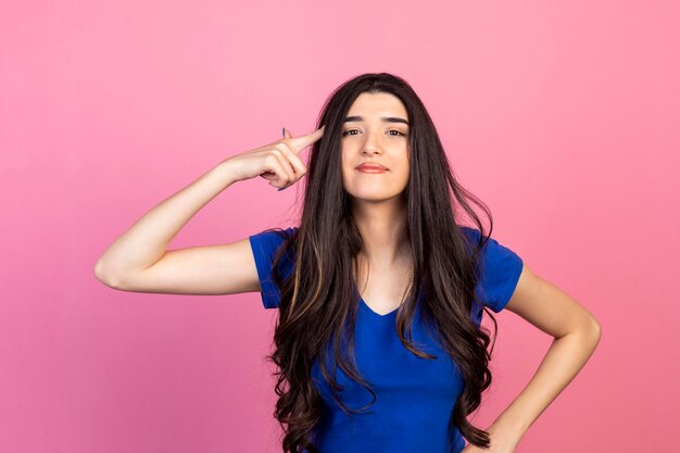 Adorable young girl points her finger to her head and looking at the camera on pink background High quality photo