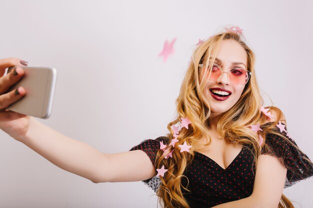 Adorable young girl with blonde long curly hair taking selfie at party, smiling, covered with pink stars confetti. Wearing colorful glasses, black dress. 