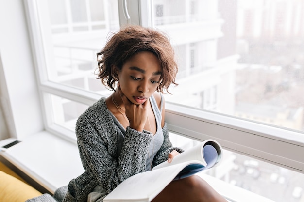 Free photo adorable young girl with short curly hair absorbedly reading magazine, book sitting on windowsill, big white window. she wearing gray cardigan, sweater, shirt.