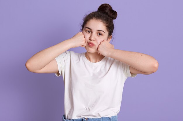 ライラックの壁に孤立したポーズをしながら彼女の頬に彼女の拳を維持する白いtシャツを着ている愛らしい若い女性