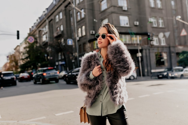 Foto gratuita adorabile giovane donna con la borsa che indossa pelliccia e occhiali da sole scuri in posa sulla città
