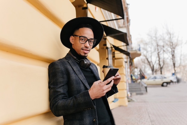 Adorable young man with brown skin standing near old building with cup of coffee. trendy african male model in casual attire holding phone while posing on the street.