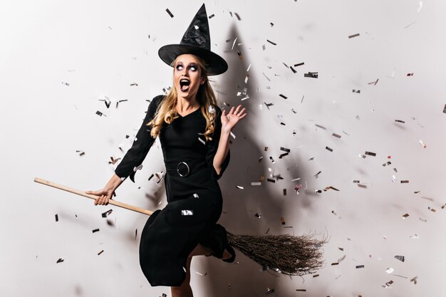 Adorable young witch sitting on broom. amazing girl in halloween hat posing on white wall.