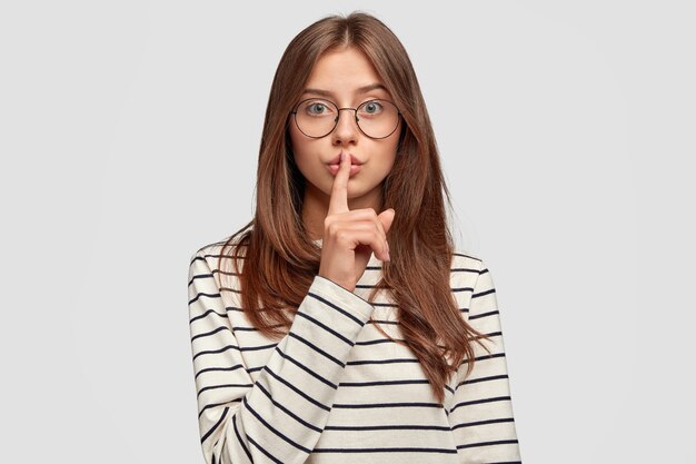 カジュアルな服を着た愛らしい若い女性は、沈黙のジェスチャーをし、直接見て、人差し指で唇に触れ、秘密にしようとし、白い壁に隔離されます。人と陰謀の概念