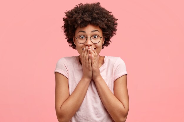 Adorable young woman with Afro haircut, has surprised glad expression, covers mouth with both palms, finds out result of entrance exam, dressed in casual t-shirt. Monochrome. Happiness