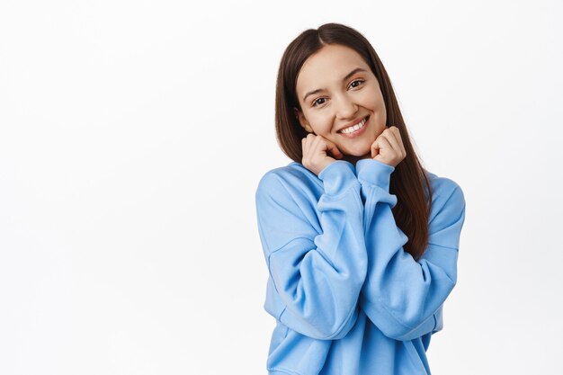 Adorabile giovane donna con un sorriso felice civettuolo, inclinare la testa, toccarsi il viso e arrossire, flirtare, guardare e contemplare un bellissimo muro bianco.