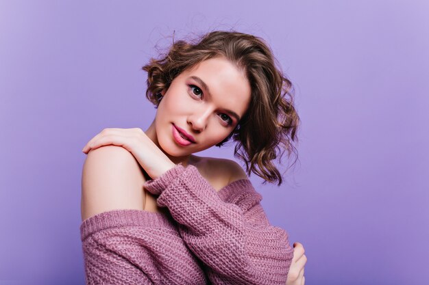Adorable young woman with dark hair looking with interest while posing in knitted sweater. attractive girl with trendy hairstyle isolated on purple wall.