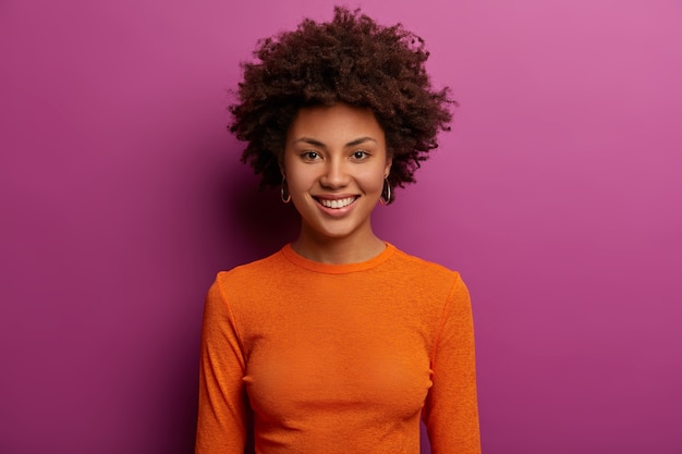 Free photo adorable young woman with natural beauty, pleasant smile, loooks happily, smiles gently, wears orange poloneck, has curly bushy hair, isolated over purple wall. pleasant emotions concept