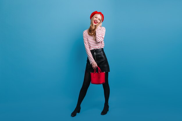 Adorable young woman with red handbag.  good-humoured french lady standing on blue wall.