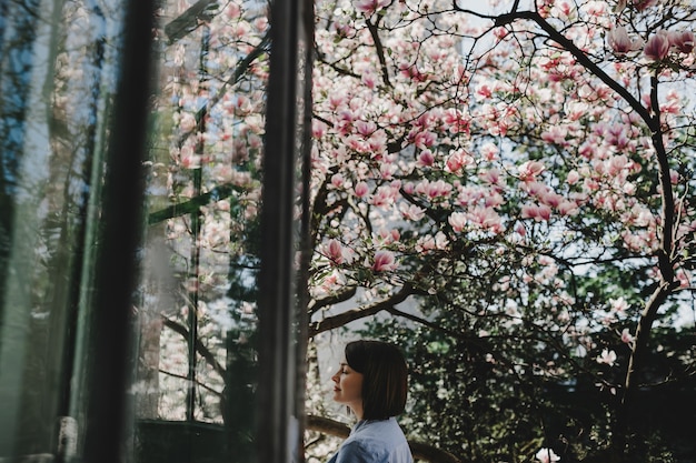 Adorable young woman with short hair stands under blooming pink tree