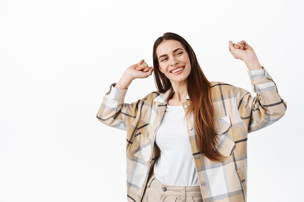 체크 셔츠를 입은 성인 미녀는 재미, 춤, 축하, 미소, 근심 없는 표정, 댄스 플로어에서 휴식, 흰 벽 위에 서 있는