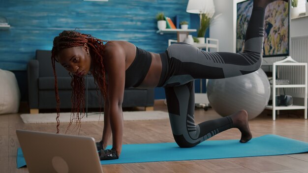 Взрослая черная женщина сидит на карте йоги и смотрит онлайн-фитнес-тренировку