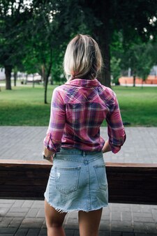Adult blonde womans back with face looking away from camera standing in park with hands in pockets w...