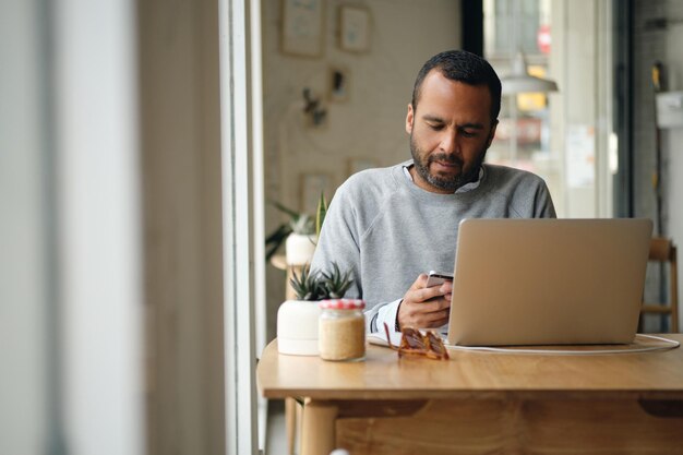 Adult businessman in sweater intently using smartphone while working on laptop in city cafe