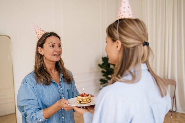 Adult caucasian cute blonde lady gives her friend small cake with candle in light room. Birthday concept