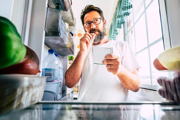 Взрослый кавказский мужчина принимает к сведению список продуктов, заглядывая в открытый холодильник дома