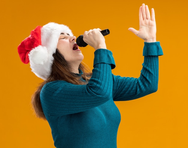 Free photo adult caucasian woman with santa hat holds mic pretending to sing isolated on orange background with copy space