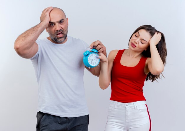 Adult couple anxious man looking and tired woman with closed eyes both holding alarm clock and putting hand on head 