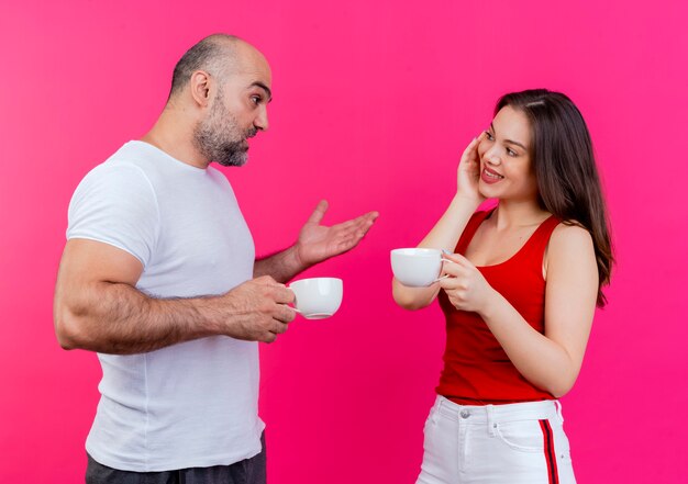 Взрослая пара, держащая чашки и смотрящая друг на друга, мужчина говорит что-то женщине и показывает пустую руку, женщина улыбается и держит руку на лице