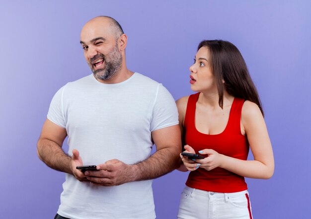 Взрослая пара, держащая мобильные телефоны, уверенный мужчина, подмигивающий, и впечатленная женщина, глядя на него